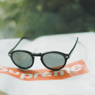 Black round sunglasses resting on a white surface with a red Supreme logo in the background.